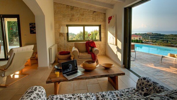 Ein modernes Wohnzimmer mit Blick auf einen Pool und das Meer, Steinwände und ein Laptop auf einem Holztisch.