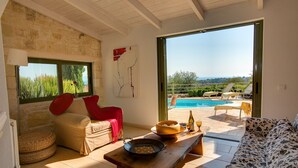 Ein Zimmer mit einer Steinsäule, einer Holzbalkendecke, einer Couch, einem Esstisch, Blick auf einen Pool und einer Tür, die nach draußen führt.