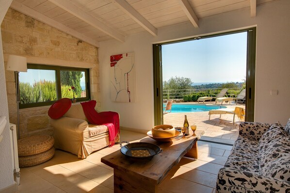 A room with a stone wall, a wooden ceiling, a sofa, a dining table, a pool view, and a door leading outside.
