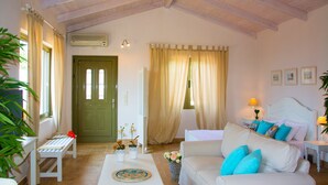 Una sala de estar acogedora con techo alto, una puerta verde, un sofá blanco con cojines azules, una mesita con un jarrón y una ventana con cortinas.
