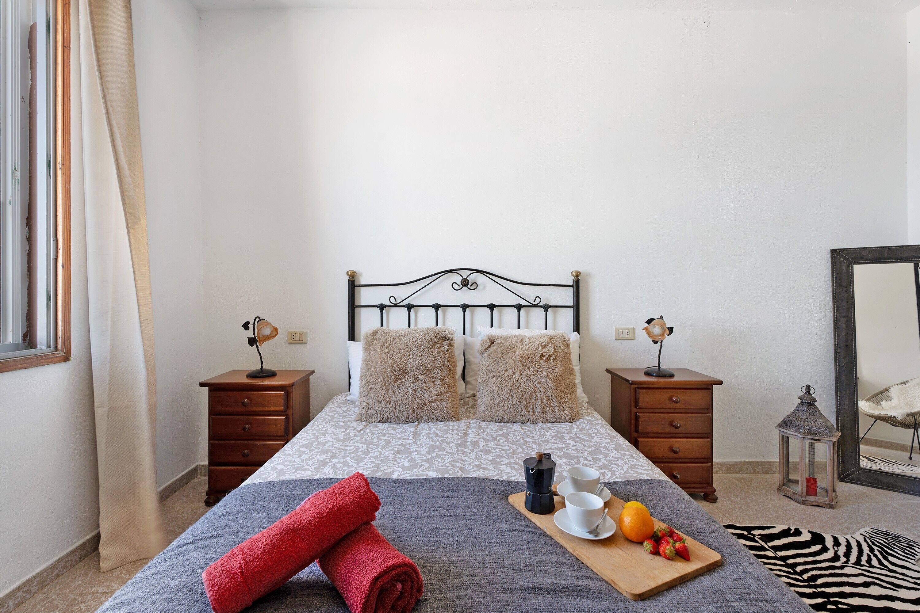 A neatly arranged bedroom with a bed, two bedside tables, a nightstand, a mirror, and a window with curtains.
