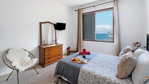 Une chambre à coucher comprenant un lit, une chaise, une commode, un téléviseur à écran plat et une fenêtre donnant sur la mer.