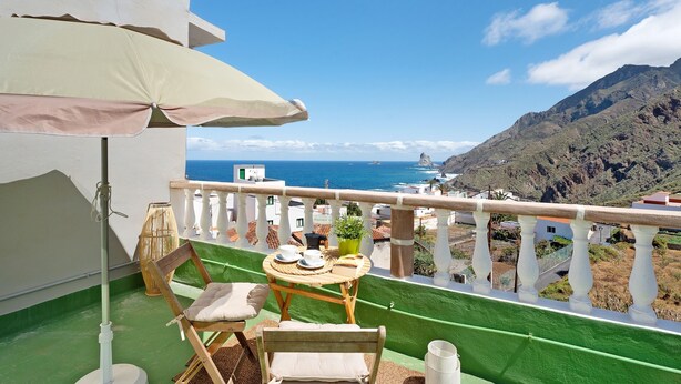 Un balcon doté d’une table, de chaises et d’un fauteuil suspendu, offrant une vue sur un paysage côtier avec des montagnes et la mer.