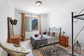 Quarto com cama, uma cadeira, cômoda, espelho, janela com cortinas e vista para montanhas.