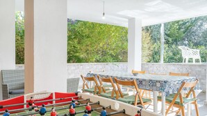 Een tafelvoetbaltafel, een terras met stoelen en tafels, en een uitzicht op bomen door een raam.