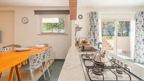 Een keuken met een gasfornuis, een eettafel met stoelen, een klok en een raam met uitzicht op de omgeving.