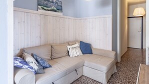 A corner living room with a beige sectional sofa, a wooden accent wall, a framed landscape painting, and a hanging light fixture.