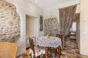 Un coin repas avec une table ronde, des chaises, une nappe décorative, un panier de fruits et un mur en pierre.