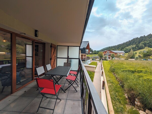 A balcony with a table and chairs, a glass door, and a view of a green landscape and buildings in the distance.