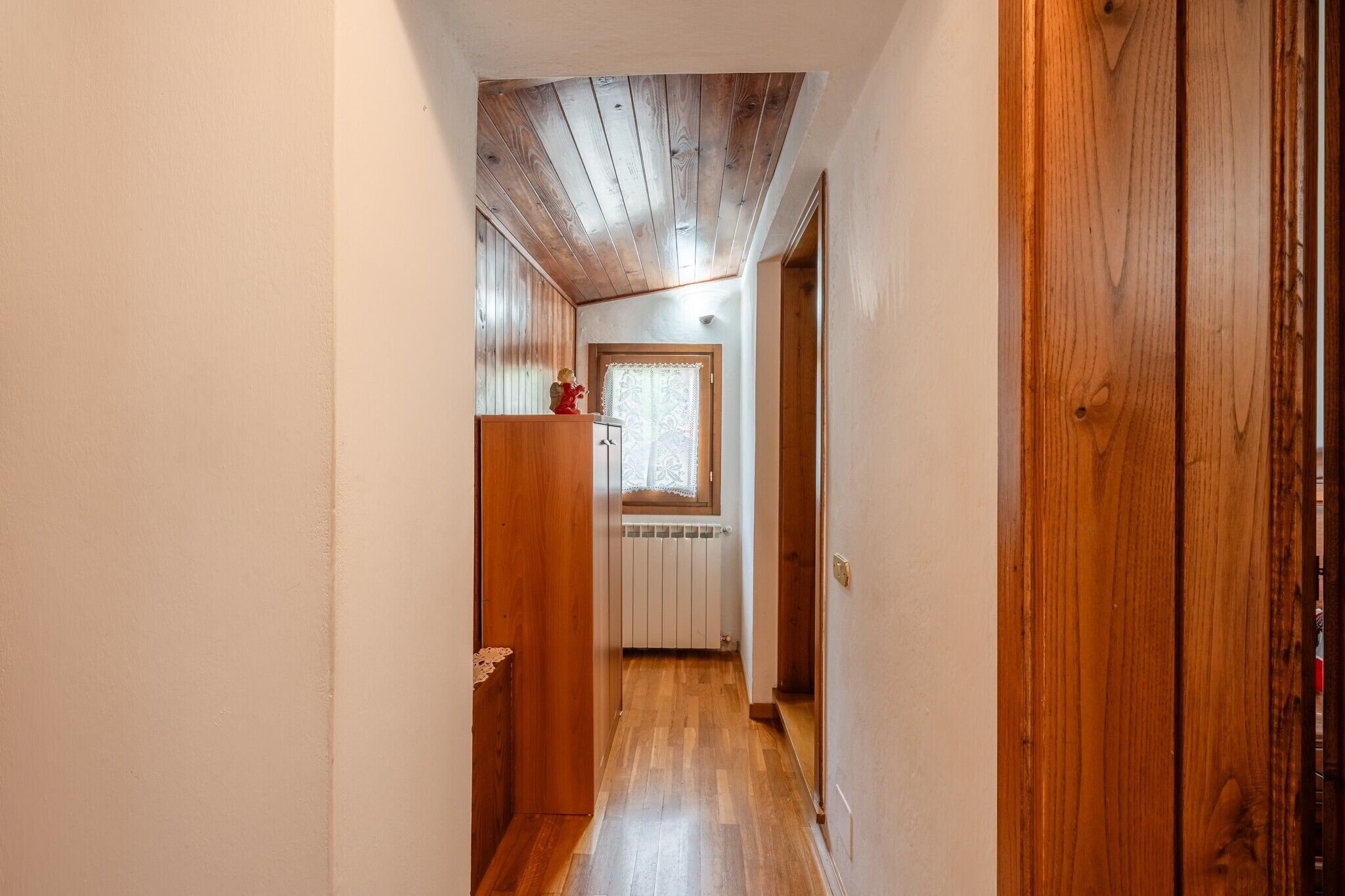 Um corredor estreito com pisos e paredes de madeira, uma porta de madeira à direita e uma janela com cortinas de renda ao longe.