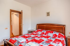 Une chambre à coucher avec un lit en bois, une couvre-lit à motifs floraux rouge et gris, une petite table de chevet et une porte donnant sur une autre pièce.