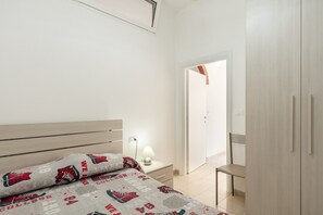 A bedroom with a bed, bedside table, lamp, chair, wardrobe, and a door leading to another room.