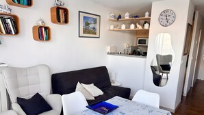 A living room with a sofa, chairs, a table with a blue folder, bookshelves with books, a framed picture, kitchen area with a microwave, and a clock on the wall. - Apartment 'Résidence La Croisette' with Sea View and Private Terrace (Agde)