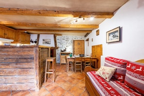 A rustic kitchen with wooden beams, a stone wall, a wooden dining table, and a red patterned sofa. - Studio Apartment "2-4 Personnes" with Mountain View, Private Terrace & Wi-Fi (BEAUFORT)