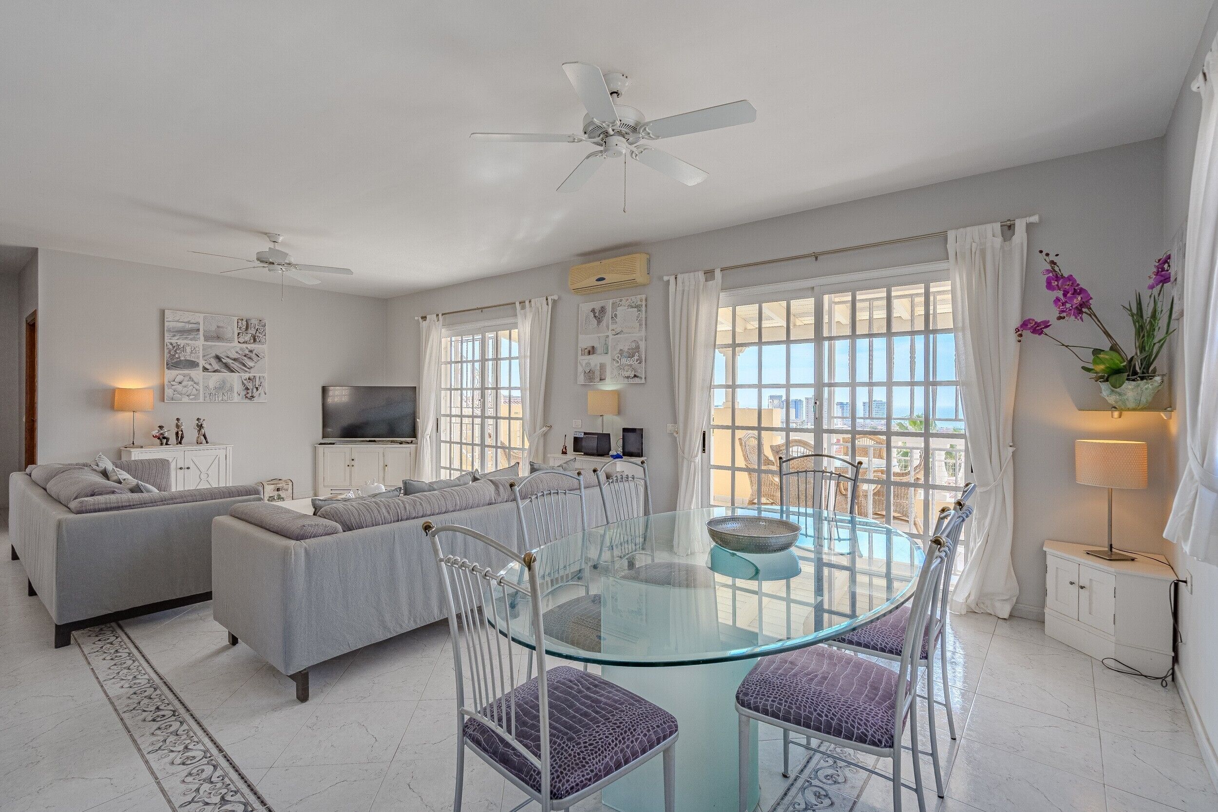 A modern living room with a glass dining table, grey sofas, and a ceiling fan.