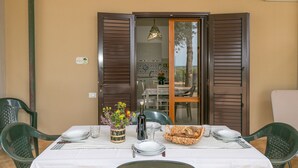 Une table à manger dressée avec des assiettes, des verres, une bouteille et un panier de pain, donnant sur l’extérieur grâce à une porte vitrée.