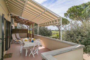 A balcony with a striped awning, a table with chairs, and a view of trees and greenery.