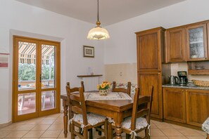 Zona pranzo con tavolo e sedie in legno, lampadario a sospensione, cucina con mobili in legno e vista attraverso una porta di vetro.