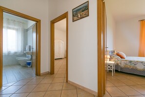 A bedroom with a bed, a nightstand, a window with curtains, a bathroom with a toilet, and a hallway leading to another room.