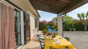 Une terrasse extérieure avec une table dressée pour deux personnes, des chaises, une pergola et une vue sur un jardin agrémenté de fleurs rouges et d’une piscine bleue.