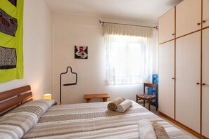 Un dormitorio con cama, mesita de noche, una silla, armario y ventana con cortinas.