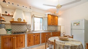 A kitchen with wooden cabinets, a dining table with chairs, a refrigerator, and a window with curtains. - Villa Margi with Sea View, Shared Pool, Private Terraces and Wi-Fi (Scopello)