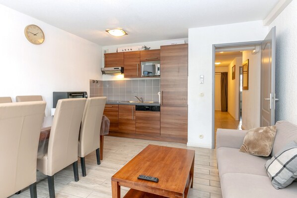 A modern kitchen with wooden cabinets, a built-in microwave, and a toaster. There is a dining area with a table and chairs, and a living area with a sofa and a coffee table.