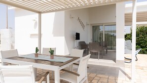 Een terras buiten met een houten tafel en witte stoelen, een pergola en een glazen deur die leidt naar een binnenruimte.