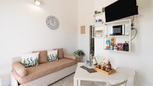 A living room with a sofa, a coffee table, a wall-mounted TV, a clock, and a shelf with kitchen items. - Holiday Home 'Villa Gioberta, Fiori' with Shared Pool, Wi-Fi and Air Conditioning (Menfi)