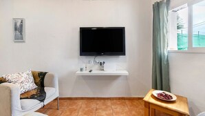 A living room with a flat-screen TV mounted on a white wall, a sofa, a small wooden table with a plate of food, and a window with curtains. - Holiday Apartment 'Casa Verol B' with Wi-Fi (Moya)