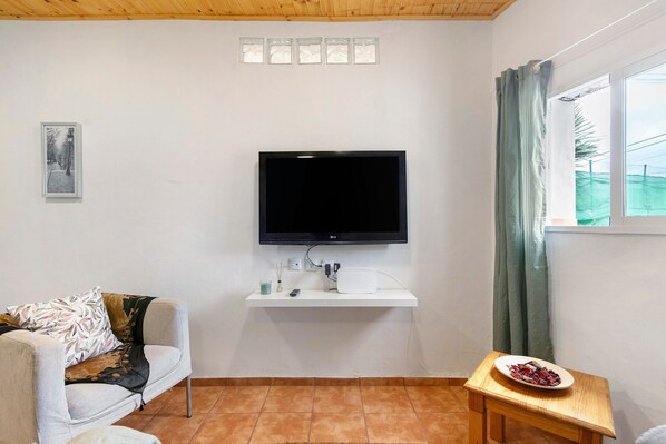 Una sala de estar con un televisor de pantalla plana montado en una pared blanca, un sofá, una mesita de madera con un plato de comida y una ventana con cortinas.