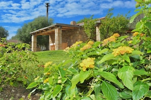 Um edifício de pedra com varanda coberta, rodeado por vegetação exuberante e plantas floridas.