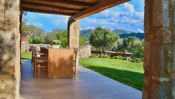 Un patio al aire libre con una mesa y sillas de madera, paredes de piedra y vistas a un paisaje con césped, árboles y montañas.