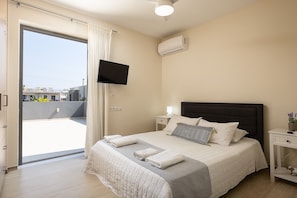 A bedroom with a bed, a TV, an air conditioner, a nightstand, and a view of the outside.