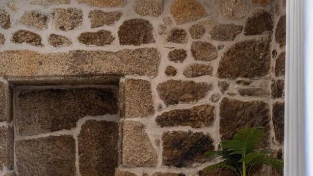 Una habitación con pared de piedra, una chimenea, una mesa redonda de madera con una vela, una planta y un banco de madera.