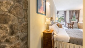Een slaapkamer met een stenen muur, een bed, een nachtkastje, een lamp, een lijst met een afbeelding, een raam met gordijnen en uitzicht op het omliggende landschap.