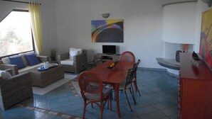 A dining area with a table and chairs, a sofa, a television, and a painting on the wall. - Villa 'Maria' with Sea View, Private Terrace and Wi-Fi (Cala Liberotto)