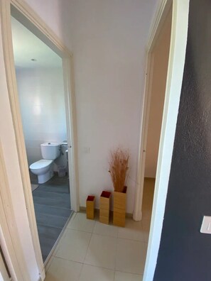 Un couloir étroit mène à une salle de bain équipée de toilettes et d’un lavabo. Deux vases en bois contenant des plantes séchées sont posés sur le sol.