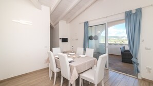 A dining area with a table set for four, white chairs, a ceiling with exposed beams, and a view through a glass door to an outdoor balcony.