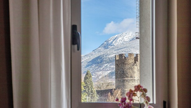 Une vue par la fenêtre sur un château, avec une montagne en arrière-plan, des rideaux à moitié tirés et un vase rempli de fleurs sur le rebord de la fenêtre.