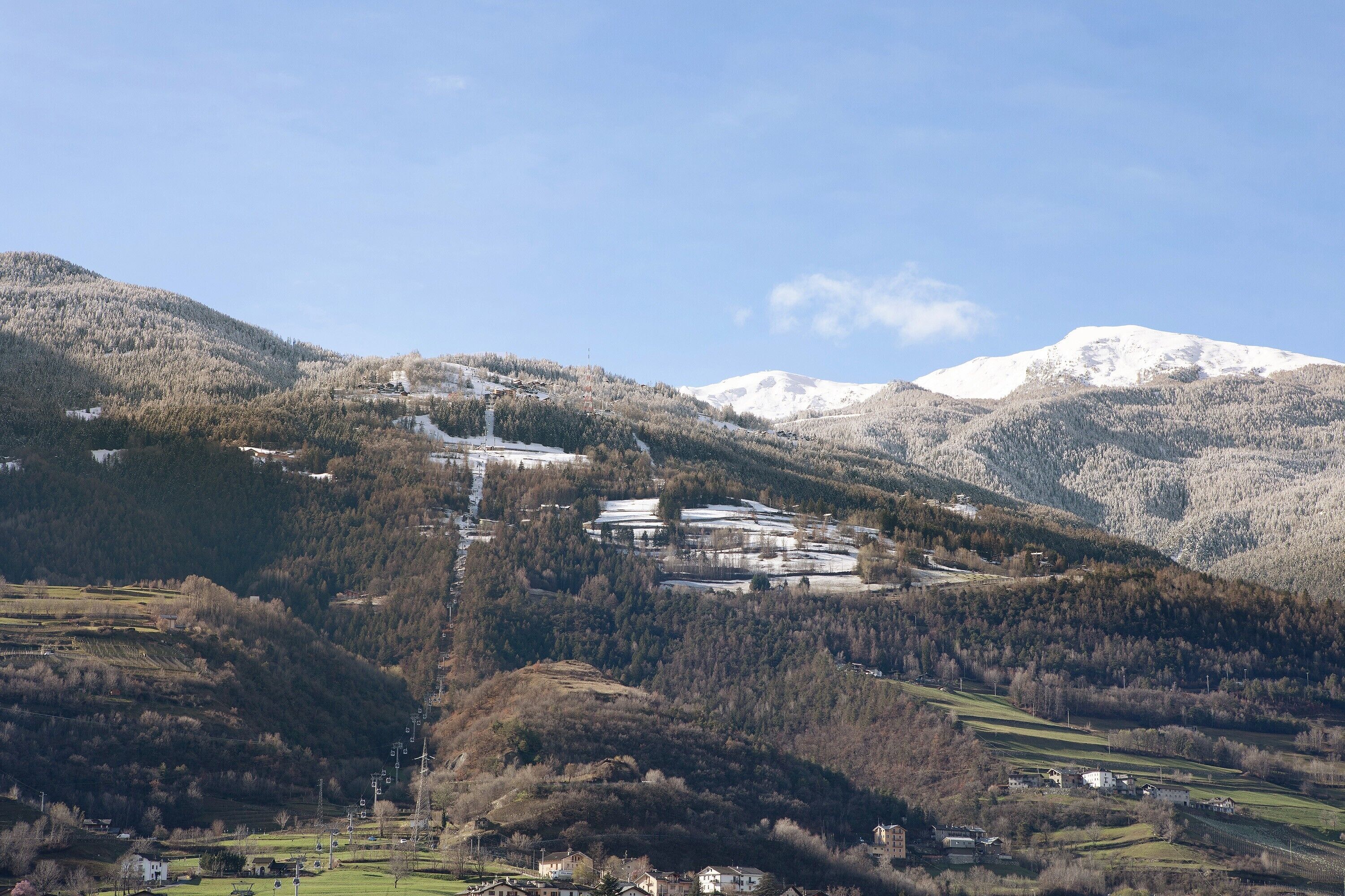 Un paysage montagneux avec un ensemble de bâtiments, peut-être un complexe hôtelier ou un chalet, entouré de collines boisées et de sommets enneigés.
