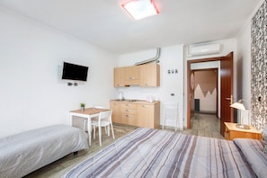 A bedroom with a bed, a desk, a chair, a TV, and a kitchenette.