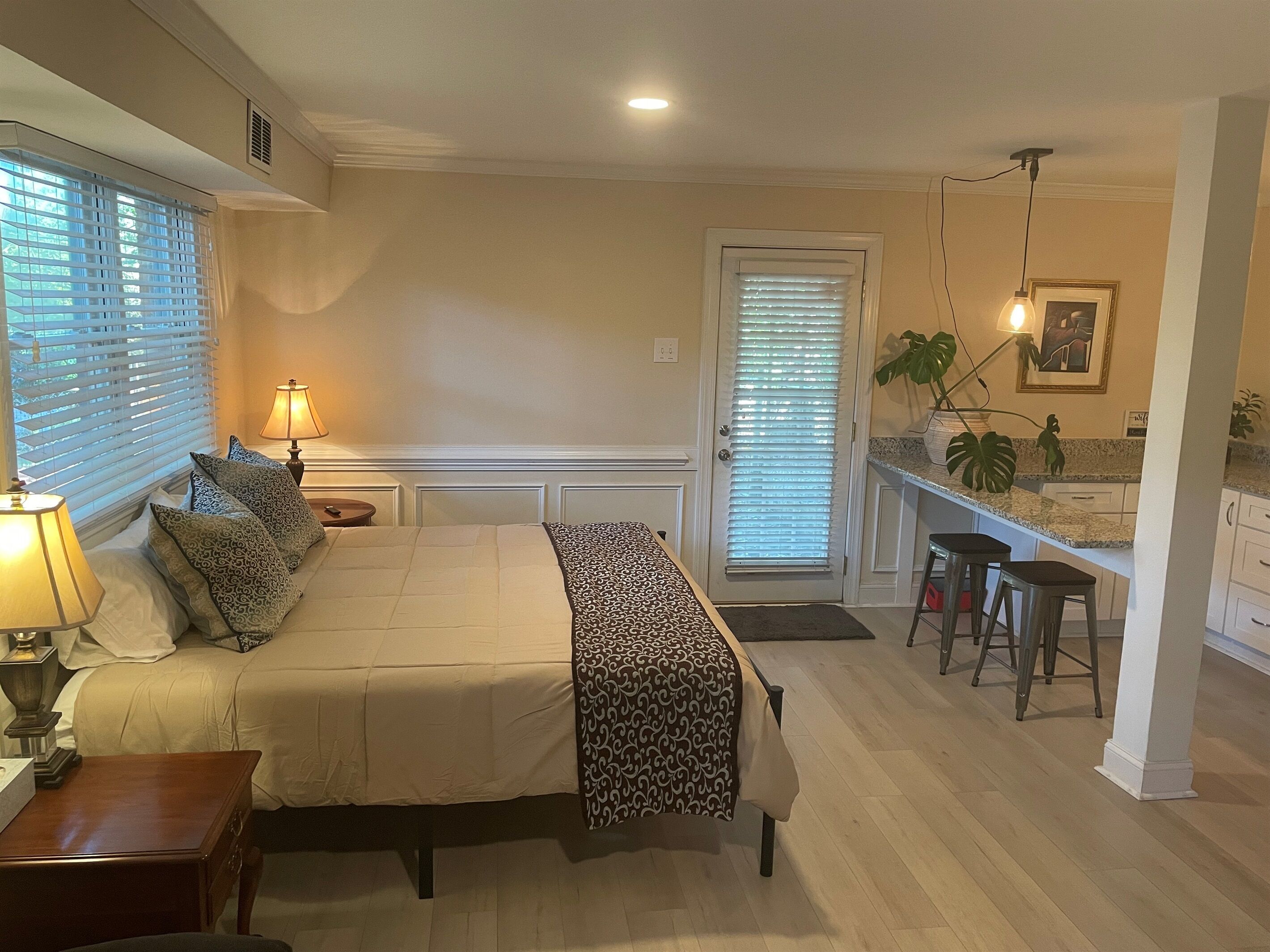 Une chambre à coucher avec un lit, des lampes de chevet, une table de chevet et un bureau. On y trouve une porte avec une fenêtre, un coin cuisine avec un plan de travail et un tabouret de bar.
