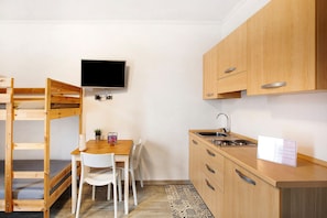 Habitación compacta con literas, una pequeña mesa de comedor con sillas, un televisor montado en la pared y una zona de cocina con armarios de madera y un lavabo.