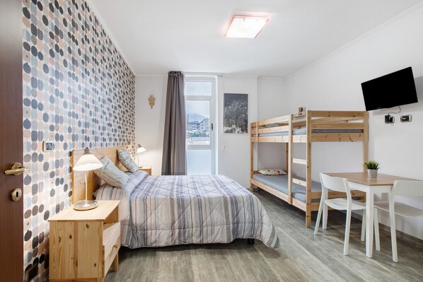 A bedroom with a bed, bedside table, lamp, bunk beds, desk, chairs, and a TV.