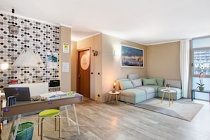 A modern living room with a sofa, a desk with a laptop, a small table with a lamp, and a wall with a geometric pattern.