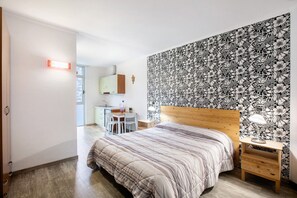 Ein Schlafzimmer mit Bett, Schreibtisch und Stuhl, ein Nachttisch aus Holz mit Lampe sowie eine Wand mit floralem Muster.