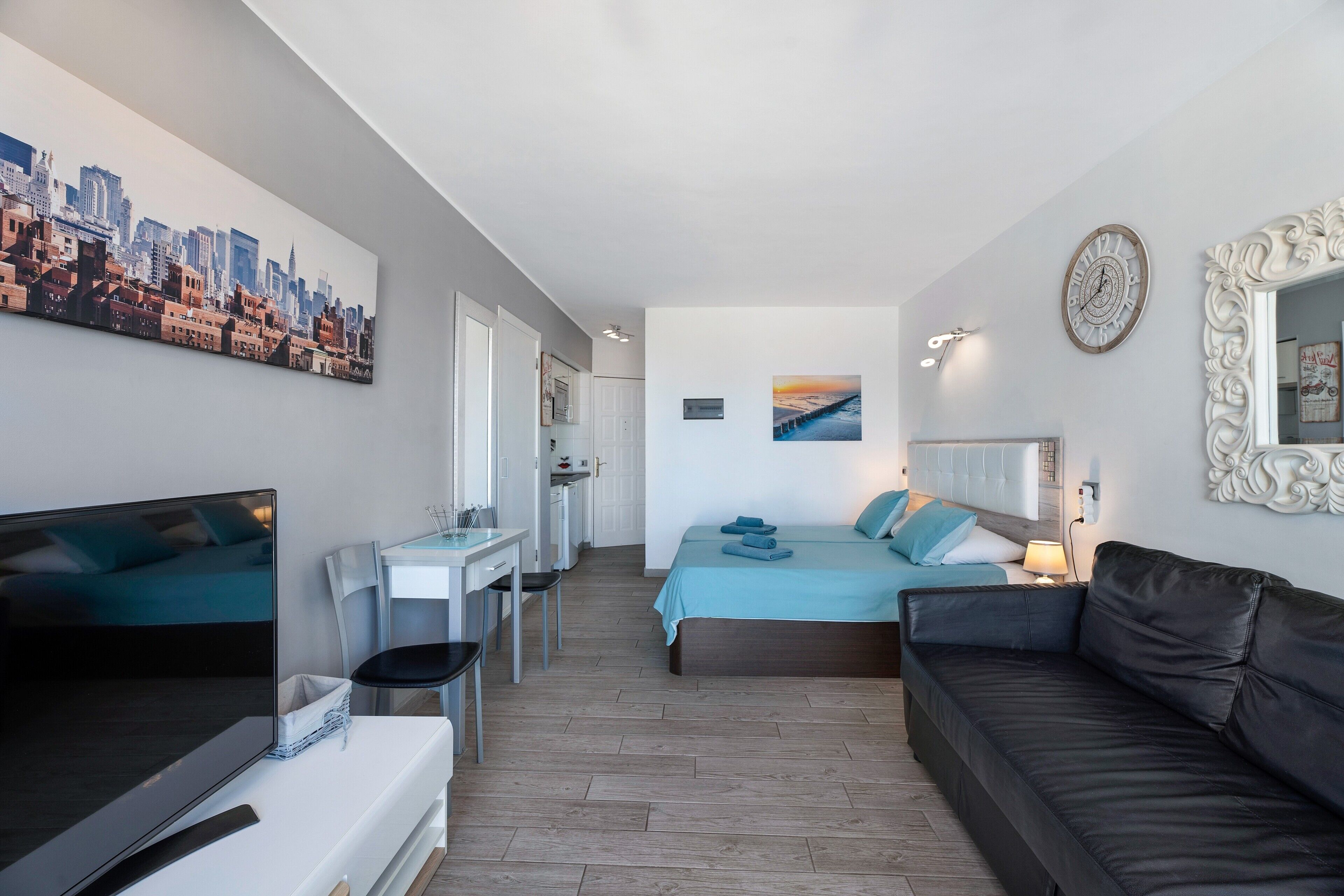 Une chambre d’hôtel moderne avec un tableau représentant un paysage urbain, un lit avec une literie bleue, un canapé en cuir noir, un bureau avec une chaise et un téléviseur.