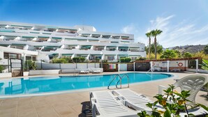 A swimming pool with lounge chairs, a fenced area, and a multi-story building with balconies. - Vacation Studio 'Las Flores 407' with Sea View, Shared Pool and Wi-Fi (Costa Adeje)