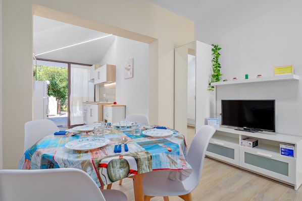 Un coin repas avec une table dressée pour quatre personnes, un téléviseur posé sur un meuble et une vue sur l’extérieur.
